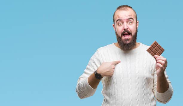 istock Young hipster man eating chocolate bar over isolated background with surprise face pointing finger to himself 1127774363