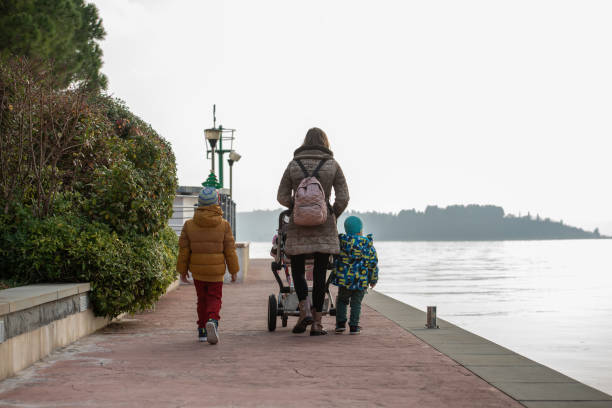 View from behind of a young mother walking on a promenade along the sea pushing a stroller and her two toddler boys walking next to her on a beautiful winter day.