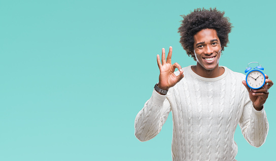 Afro american man holding vintage alarm clock over isolated background doing ok sign with fingers, excellent symbol