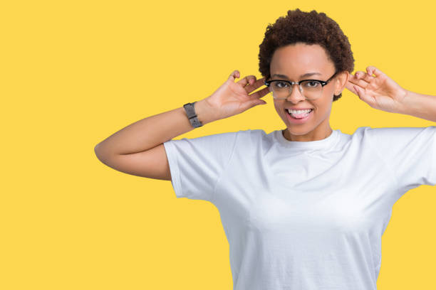 belle jeune femme afro-américaine portant des lunettes sur le fond isolé souriant tirant des oreilles avec des doigts, geste drôle. problème d'audition - casting glass photos et images de collection