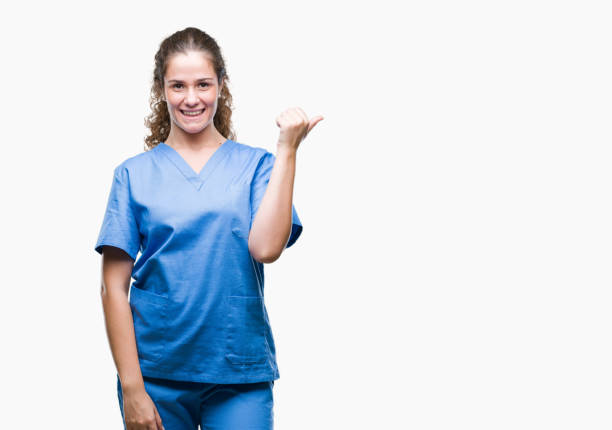 Young brunette doctor girl wearing nurse or surgeon uniform over isolated background smiling with happy face looking and pointing to the side with thumb up. Young brunette doctor girl wearing nurse or surgeon uniform over isolated background smiling with happy face looking and pointing to the side with thumb up. sign human hand pointing manual worker stock pictures, royalty-free photos & images