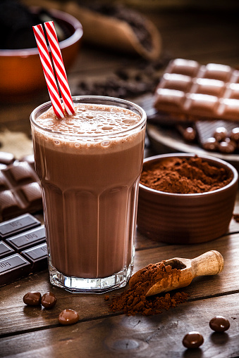 Preparing homemade chocolate milkshake. Chocolate milkshake glass shot on wooden kitchen table. The milkshake glass has two red and white drinking straws inside. A brown bowl filled with ground cocoa is beside the glass. The milkshake glass is surrounded by several chocolate bars. Predominant color is brown. Low key DSRL studio photo taken with Canon EOS 5D Mk II and Canon EF 70-200mm f/2.8L IS II USM Telephoto Zoom Lens