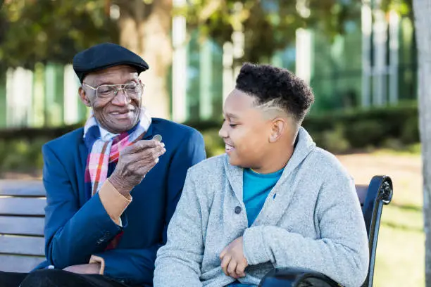 A 10 year old African-American boy sitting on a bench with his 79 year old great grandfather. They are smiling as the senior man hands his great grandson a quarter.