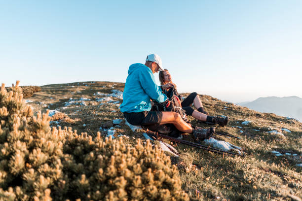 Young man and woman hiking in the mountains. Climbing up the hill with a backpack. Two adults being active. Sunny walk in the nature. Happy couple taking a break,  enjoying the view, eating brunch and talking to each other.