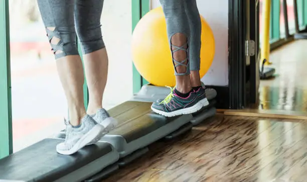 Cropped view of two women standing side by side in a gym on step exercise equipment, doing calf raises. The one on the right is a mature African-American woman in her 50s and her friend is a senior woman in her 60s.  Only their legs are visible.