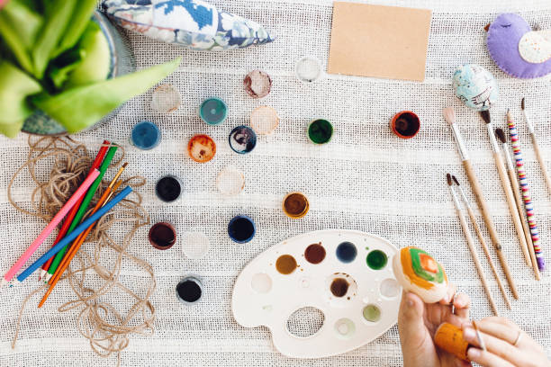 istock Top view of hands holding and painting easter egg on rustic table with paint,brushes, tulips bouquet, space for text. Child decorating easter egg, holiday preparations. Happy Easter concept 1127804092