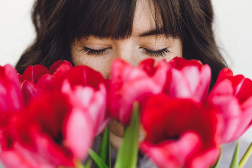 Beautiful brunette girl portrait with red tulips closeup on white background indoors, space for text. Stylish young woman  smelling tulips with closed eyes. Fresh aroma scent concept