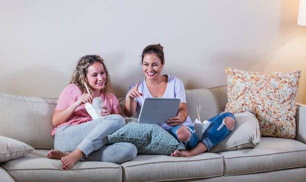 istock Hispanic sisters on sofa eating take out, digital tablet 1127805995