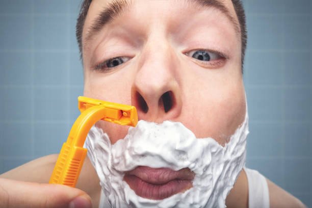 Close-up of a mans face. He is using a razor and foam to shave his beard.