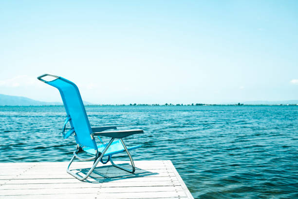 a lonely blue deck chair at the end of a weathered wooden pier next to the water, in a relaxing ambient, with a blank space on the clear sky