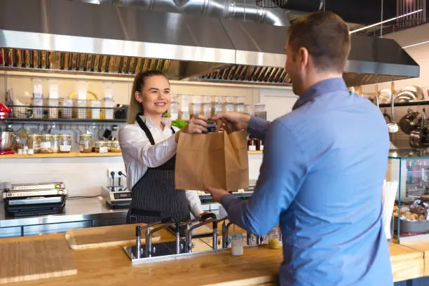 Photo of Happy waitress waring apron serving customer at counter in small family eatery restaurant. Small business and entrepreneur concept with woman owner in eatery with takeaway service delivery