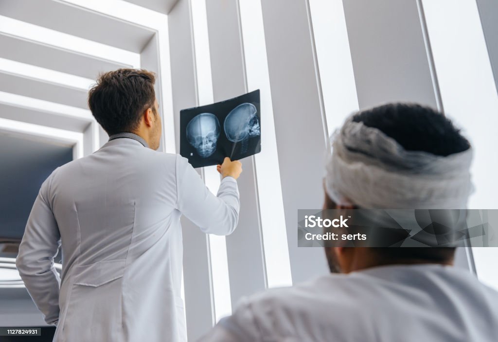 Young doctor man looking x-ray of the patient's skull Healthcare professional young doctor examining and  analyzing x-ray CT Scan test of human head results of the patients skull with his patient in a medical hospital clinic. Physical Injury Stock Photo