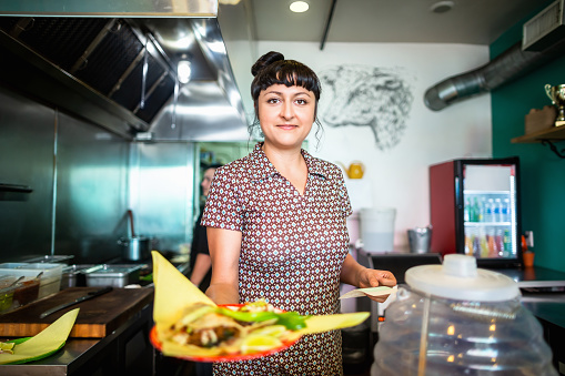 A woman standing behind the counter in a colorful taqueria holds a plate of food out to the camera.