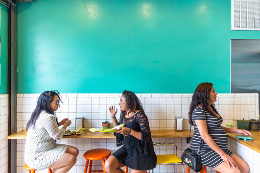 Two young women are sitting at the bar, chatting and eating tacos, in a Mexican restaurant. behind them a third woman, who is pregnant, is sitting at the counter talking to someone outside the frame.