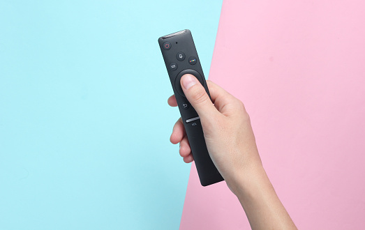 Female hand holds the TV remote on a pink  blue pastel background. Top View