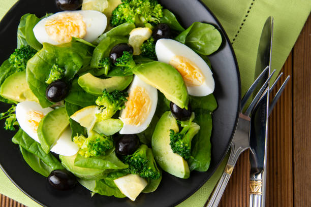 Avocado salad with broccoli, spinach, olives and boiled eggs in black plate, over wooden table. Healthy food. Avocado salad recipe. Avocado salad with broccoli, spinach, olives and boiled eggs in black plate, over wooden table. Healthy food. salad fruit lettuce spring stock pictures, royalty-free photos & images