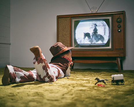 A period correct 1960's television displaying a cowboy scene on the screen (not simulated) with a little boy dressed as a cowboy watching the screen. Image toned to match the era.