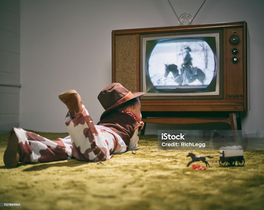 Vintage TV and Little Boy Cowboy A period correct 1960's television displaying a cowboy scene on the screen (not simulated) with a little boy dressed as a cowboy watching the screen. Image toned to match the era. Television Set Stock Photo