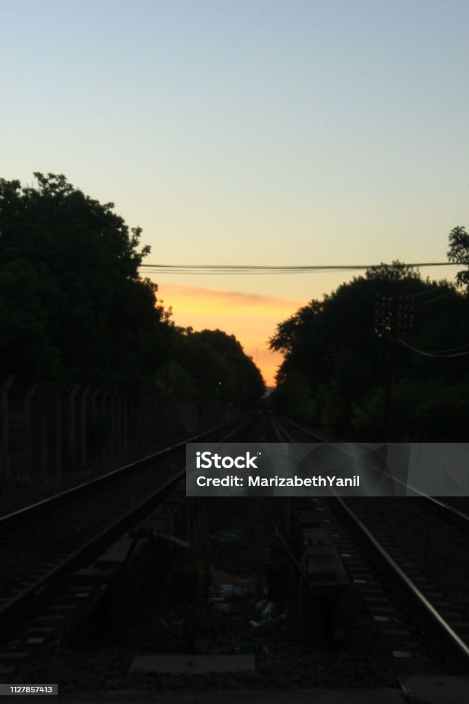 VIEWS OF TRAIN TRACK IN FRONT WITH SUNSET IN THE BACKGROUND FRONT VIEW OF TRAIN TRACKS, WITH TREES ON THE SIDES, WITH CLEAR SKY IN THE BACKGROUND, AT SUNSET Argentina Stock Photo