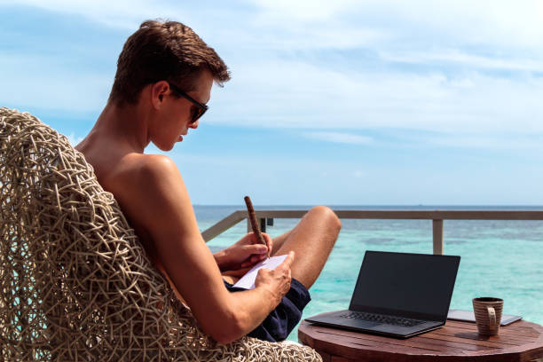 istock young man in swimsuit working on a laptop in a tropical destination. Taking notes on a notebook 1127857643