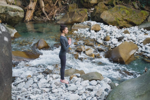 istock Young slim woman practicing yoga outdoors on stone shore of mountain river. Unity with nature concept. Girl standing in profile with prayer hands 1127865603