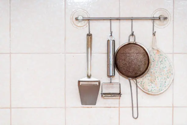 Kitchen utensils hanging on wall with sieve, metal peeler and towel mounted with suction cups closeup of tiled wall