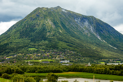A majestic rocky mountain in Norway are captured on a cloudy summer day. The mountain slopes are overgrown with trees, and at the bottom of the mountain there are a settlement and a body of water.