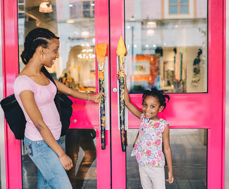 Little girl and her mother hold the handles of a retail shop about to go in, maybe on a mother daughter bonding trip doing some shopping. Candid cute moment between mother and daughter. Family is African American and child is 4 years old. Having fun and browsing shops
