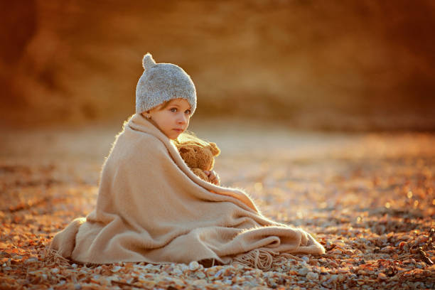 Side view of little girl in hat cuddling in warm plaid with toy while sitting on beach in sunshine