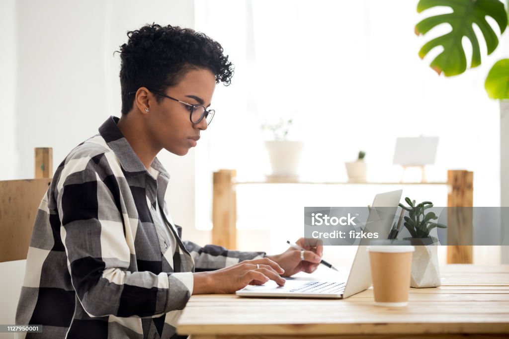 African american young woman using laptop working or studying online African american young woman using laptop working studying online, serious black girl student worker looking at laptop making notes typing busy on computer research, search information in internet Writing - Activity Stock Photo