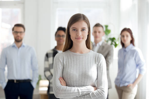 Female leader smiling looking at camera with team at background Confident young female leader, office worker, intern or company employee smiling looking at camera with team at background, happy successful businesswoman professional manager business coach portrait reduction looking at camera finance business stock pictures, royalty-free photos & images