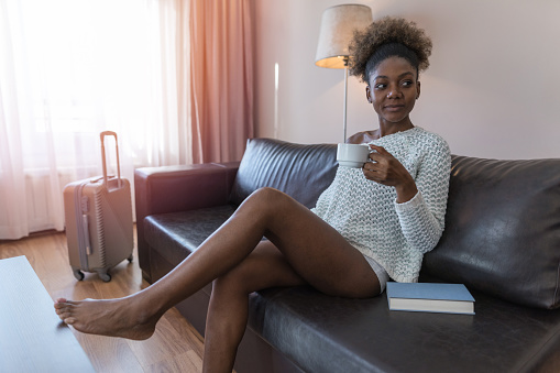 Attractive African-American woman resting in her hotel room, drinking coffee and reading book