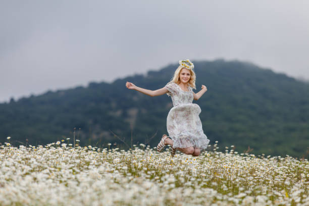 Slim girl in light dress jumping in chamomile field Attractive young woman in camomile wreath hopping with arms outstretched in daisies field in overcast day gladiator shoe stock pictures, royalty-free photos & images