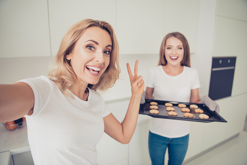 Close up photo showing v-sign two people mum and teen daughter enjoy spending time make take selfies hot heart shape figure cookies housewife help wear white t-shirts jeans in bright flat kitchen