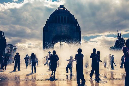 Mexico City, Mexico, June 5, 2011 - The silhouette of dozens of children and teenagers play with water from the fountains installed on the floor in front of the Revolution Monument (the Mexican National Monument), in the center of Mexico City, near Paseo della Reforma. The monument, completed in 1938 in art deco and surrounded by gardens and open spaces, is visited every year by thousands of tourists and Mexican citizens.