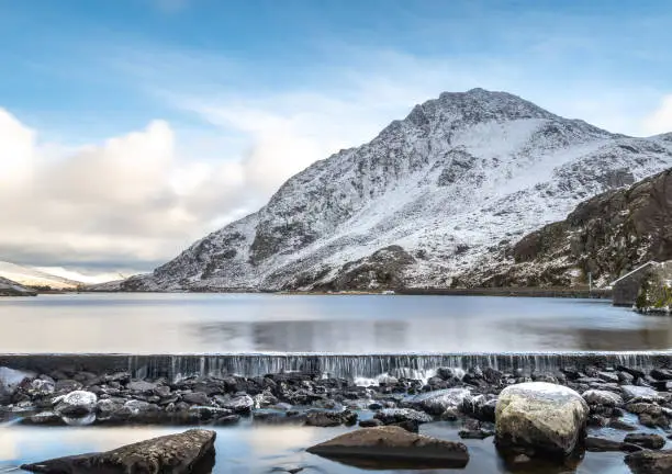 water flowing over the weir from Llyn Ogwen looking over the wintry lake towards Tryfan covered in snow