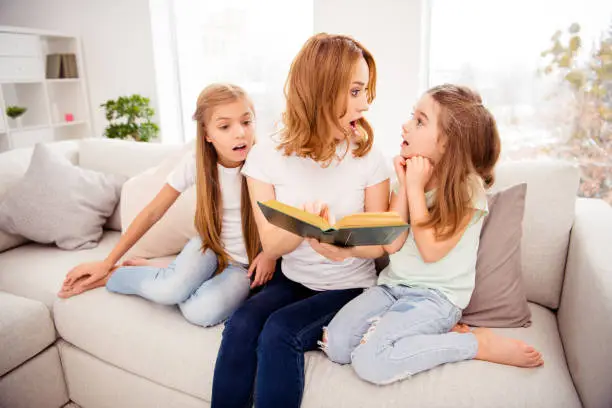 Portrait of nice cute amazed shocked lovely attractive charming cheerful cheery redhair people mom mommy mum reading book pre-teen girls sitting on divan in house indoors