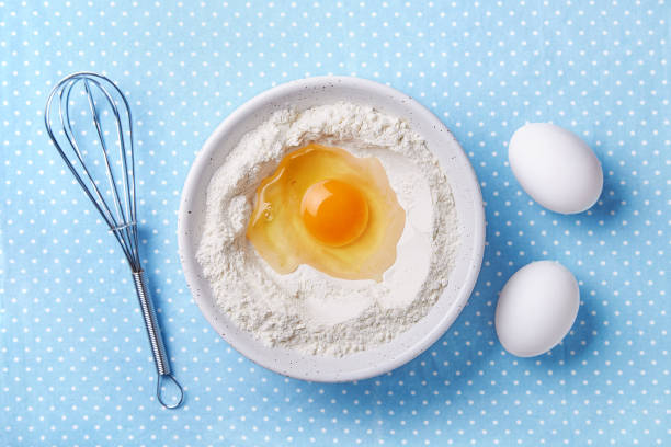 Broken egg yolk and flour in a bowl, whisk and white eggs on the side. Food preparation concept. Baking ingredients. Top view stock photo