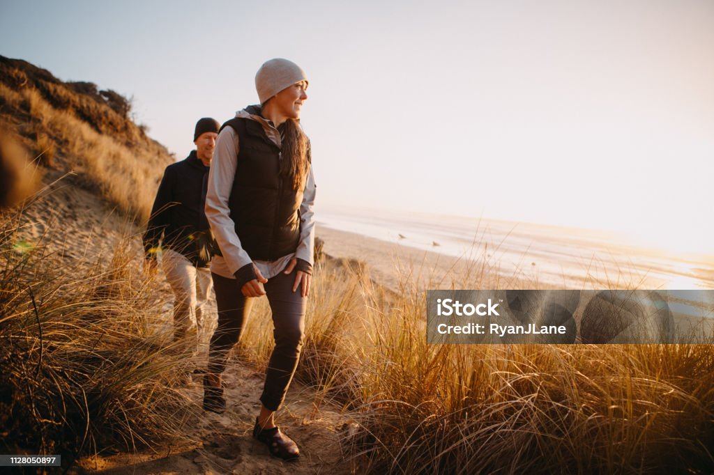Loving Mature Couple Hiking At Oregon Coast A senior couple explores a beach in Oregon state, enjoying the beauty of sunset on the Pacific Northwest coast.  They hike up a sand dune, the ocean visible in the background. Walking Stock Photo