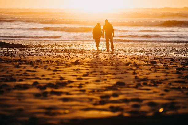 A senior couple explores a beach in Oregon state, enjoying the beauty of sunset on the Pacific Northwest coast.  They walk hand in hand, the ocean visible behind them.