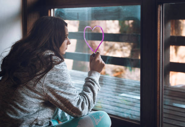 Millennial girl draws a heart with pink lipstick on a window.