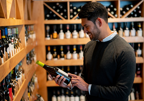 Handsome man at a wine store reading the label on bottle smiling very happy