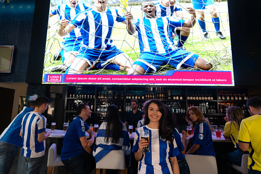 Happy female football fan at watching a game at a sports bar and drinking beer while looking at the camera smiling
