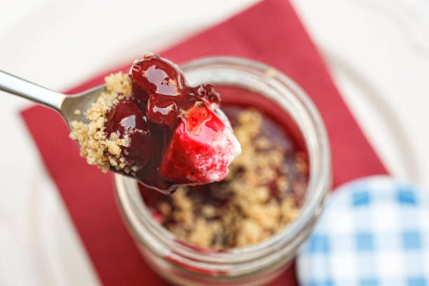 Spoonful of sour cherry cheese cake in a glass jar with biscuit crumbs Spoonful Panna Cotta Cheesecake with sour cherries served in a glass jar with biscuit crumbs - top view cake jar stock pictures, royalty-free photos & images