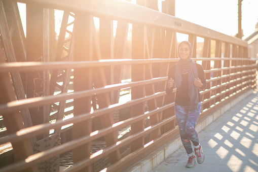 A Muslim woman is jogging on a bridge on a sunny day. She is wearing a head scarf.