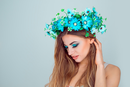 Young beautiful woman with floral crown on head in side profile looking down posing on light green blue studio background with copy space Multicultural beauty model with headband from anemone flowers