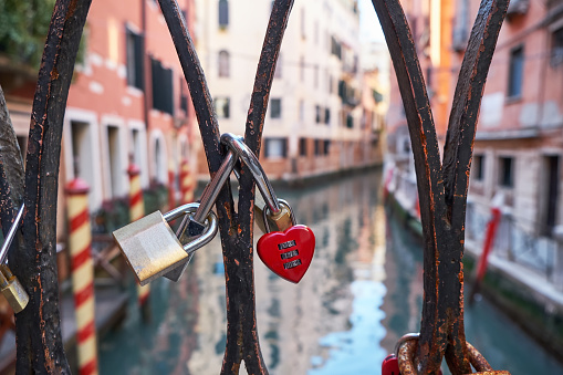 Combination red heart-shaped and other padlocks on the bridge in Venice, Italy. Sunny day, historical buildings and the canal in the background. Close up. Valentine's Day concept.