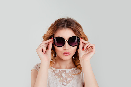 Pretty woman in round sunglasses smiling isolated light grey gray background holding her glasses puckering lips about to send a virtual kiss.