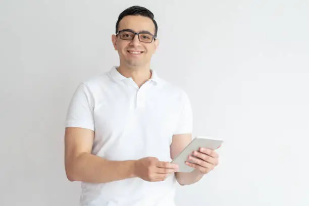 Smiling young man using tablet computer. Attractive guy wearing glasses, browsing on tablet and looking at camera. Technology concept. Isolated front view on white background.