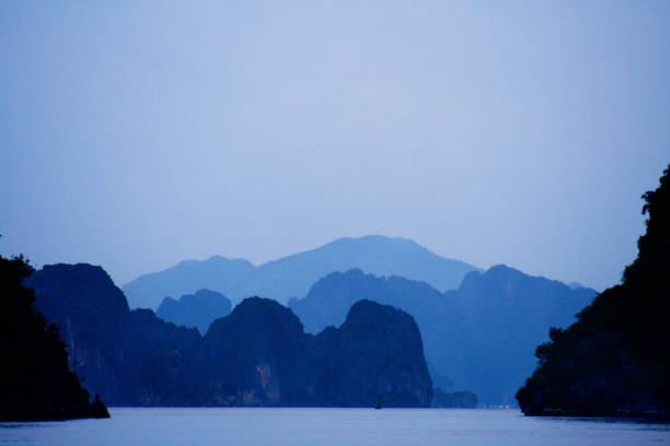 Bright orange sunlight pierces through the picturesque limestone cliffs over Halong Bay, reflecting softly on the serene waters, accompanied by breathtaking hues of orange and blue across the sky.
